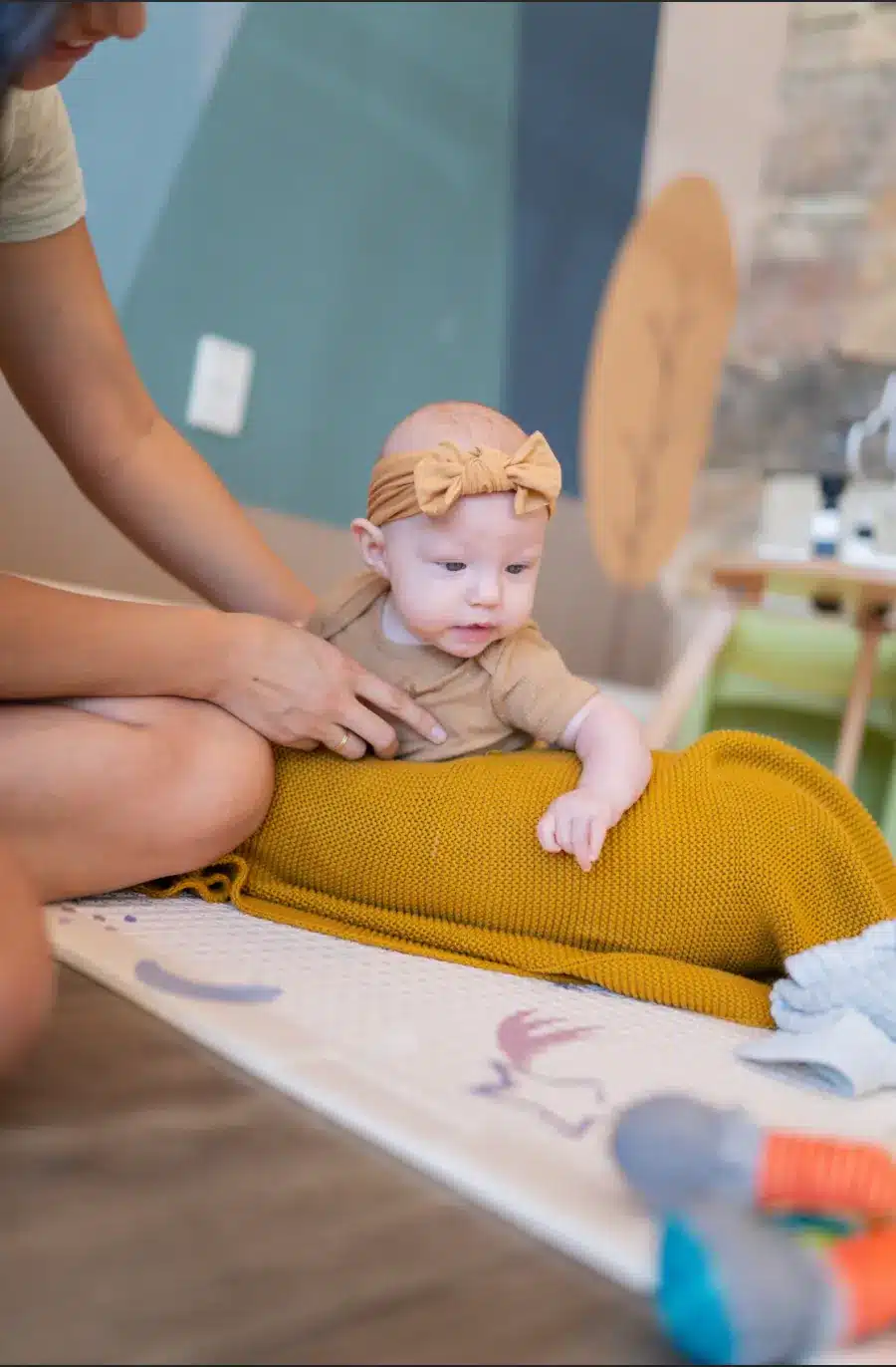 Smiling baby in tan onesie and headband sits on a mustard-yellow blanket, supported by a person.
